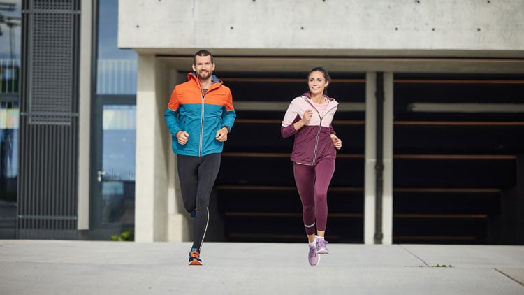 HERVIS Laufberater Zwei Personen joggen nebeneinander auf Beton vor einem modernen Gebäude mit großen Fenstern; links eine Person in blau-orangefarbener Jacke und schwarzen Leggings, rechts eine in bordeaux-pinker Jacke und passenden Leggings