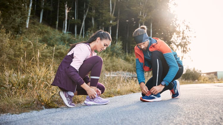 Laufschuhe Kaufberatung Zwei Personen binden sich auf einem gepflasterten Weg in einer natürlichen Umgebung die Schnürsenkel. Die Person links trägt lila Sportkleidung und helllilafarbene Schuhe, die Person rechts eine blau-orange Jacke, schwarze Hose und dunkle Schuhe. Im Hintergrund sind Bäume und Vegetation zu sehen