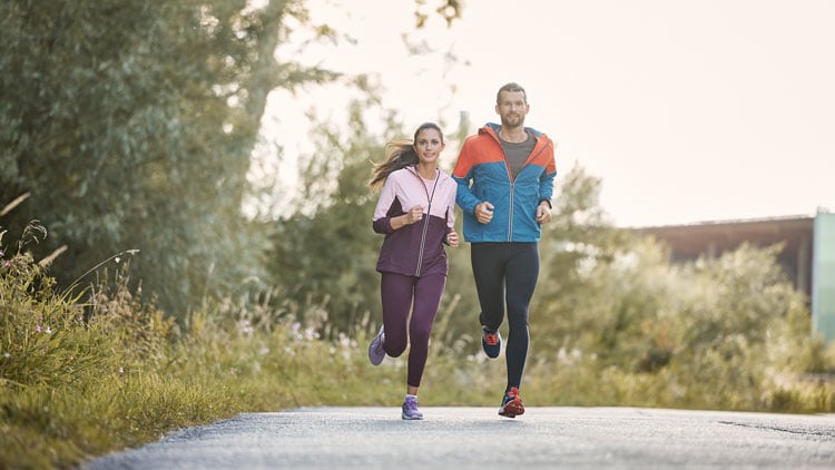 Hervis Laufberater - Laufbekleidung Mann und Frau joggen in sportlicher Kleidung durch eine grüne, bewachsene Landschaft.